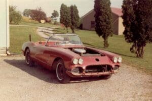 Vintage red convertible car missing a front bumper, parked on a gravel driveway