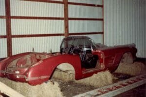 A disassembled red vintage car shell rests on hay bales in a dimly lit garage. The missing parts give a sense of neglect and nostalgia.