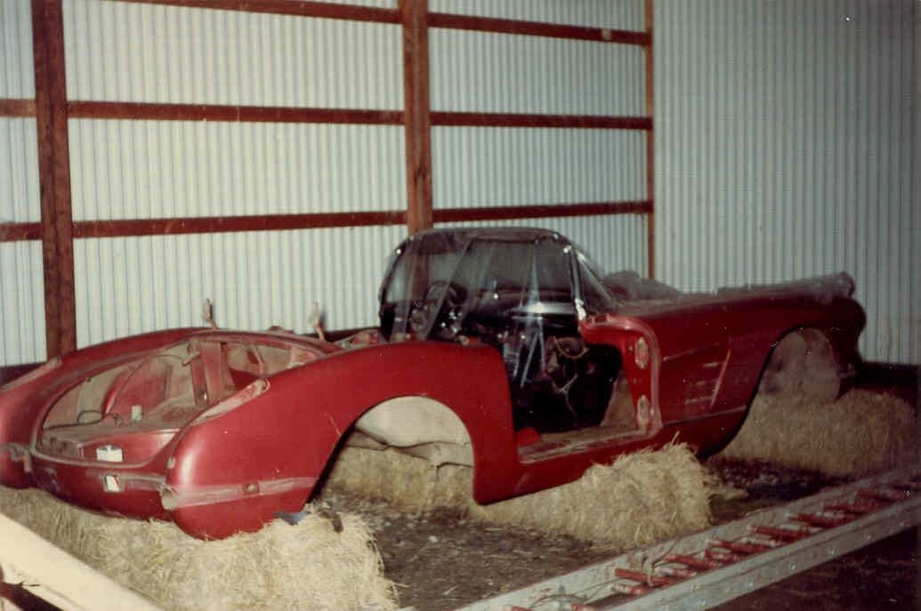 A disassembled red vintage car shell rests on hay bales in a dimly lit garage. The missing parts give a sense of neglect and nostalgia.