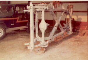 A rusted, disassembled car frame is set on the floor of a garage. A vintage Jeep with a distinctive zigzag stripe is parked beside it.