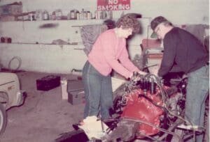 Two people work intently on a red engine in a cluttered garage. The "No Smoking" sign and various tools highlight a focused, collaborative atmosphere.