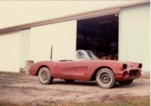 A weathered red sports car with missing parts is parked on a concrete surface outside a large open garage. The scene conveys a vintage, restoration atmosphere.
