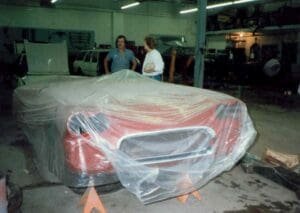 A red car in a garage, partially covered with plastic sheeting, is on jack stands. Two people converse nearby, amidst workshop tools and vehicles.