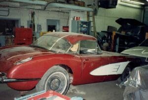 A dusty red vintage car with a white side detail is parked in a dimly lit garage. The setting has an industrial feel, with tools and equipment visible.