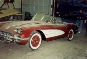 A vintage red and white convertible car parked in a garage. The car's chrome grille and round headlights stand out. A person is visible in the background.