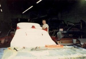 Man working in dimly lit garage, standing beside unfinished fiberglass car. Tools and automotive parts scattered around, conveying a focused, industrious mood.