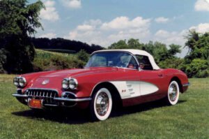 A classic red convertible sports car with white accents on lush grass, set against a backdrop of trees and a blue sky with fluffy clouds, exuding nostalgia.