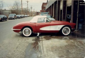 A classic red and white convertible car with a white roof, parked outside an auto garage. The street is lined with parked cars. The scene feels vintage and nostalgic.